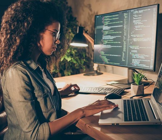 A woman sits at a desk, working on a laptop and a computer, focused on her tasks.