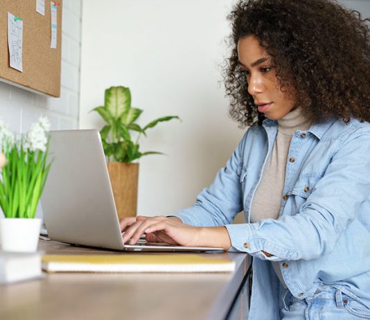 A woman with curly hair sits at a desk, focused on her laptop while working or studying.