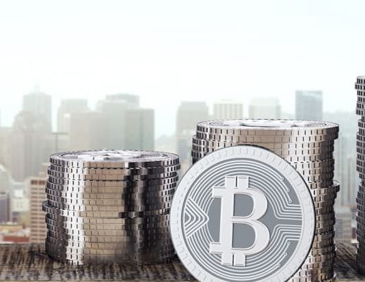 A collection of bitcoin and coins rests on a wooden table, with a cityscape visible in the background.