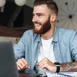 A bearded man with tattoos sits at a table, focused on his laptop, creating a relaxed and engaged atmosphere.