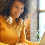 A woman with curly hair sits at a table, focused on her laptop, engaged in work or study.