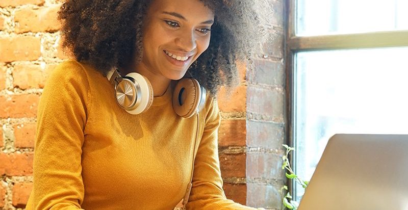 A woman with curly hair sits at a table, focused on her laptop, engaged in work or study.